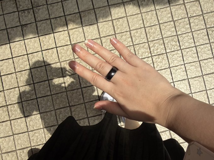A hand rests against a black garment over a tiled pavement, displaying a smooth black ring on the middle finger, with manicured nails and a pink accessory visible in the corner.