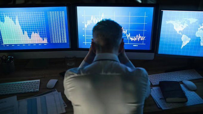A man sits with his back to the camera, looking at three computer monitors displaying financial charts, graphs, and a world map.