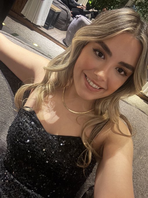 A selfie of a smiling young woman with long blonde hair wearing a sparkly black sequined top and a gold chain necklace, seated in what appears to be an upscale outdoor lounge area.