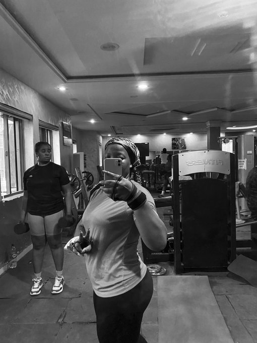 A black and white photo of two women in a gym, with one taking a mirror selfie while the other holds dumbbells in the background.