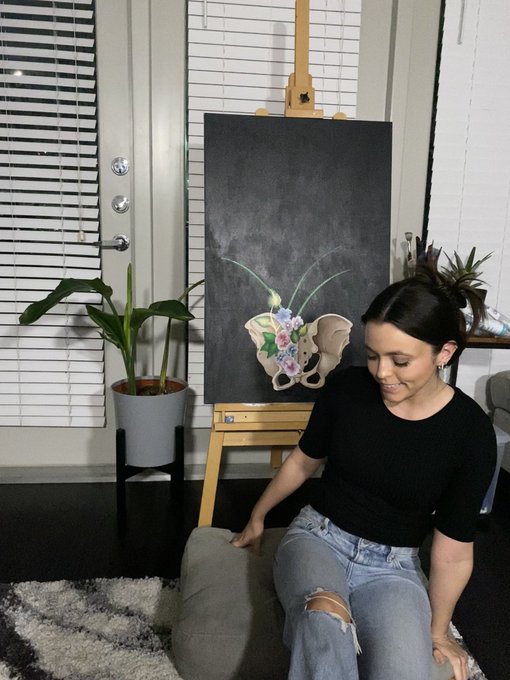 A young woman in a black top and ripped jeans sitting on a stool in an art studio, with a floral and moth-wing painting on an easel behind her.