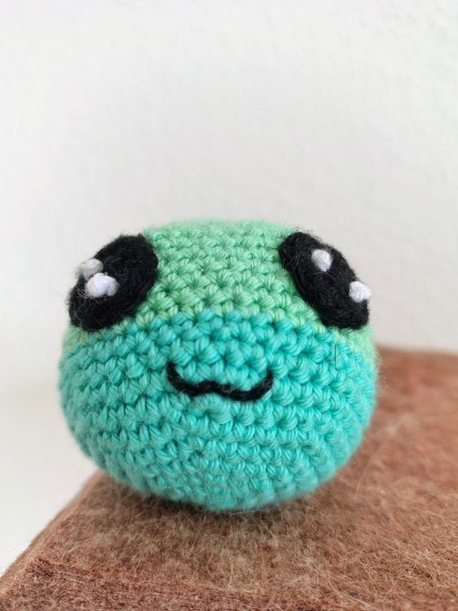 A small, round crocheted green plush toy with a smiling face and large black eyes sits on a brown surface.