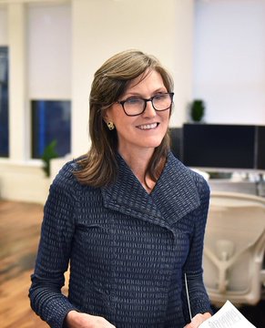 Cathie Wood, the founder and CEO of ARK Invest, is shown smiling in what appears to be a modern office setting. The image accompanies a post highlighting her bold prediction urging investors to shift from gold to Bitcoin, with her forecast that BTC could reach $1.5 million by 2030.