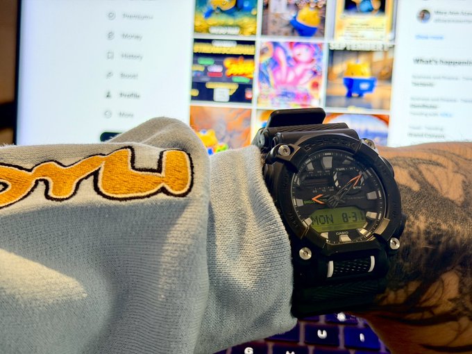 A wrist wearing a black rugged watch displaying "MON 8:31" rests against a computer monitor showing a webpage with "Music" and "Royalty" menu options, visually demonstrating the caption's claim that the watch stays in EST around the clock.