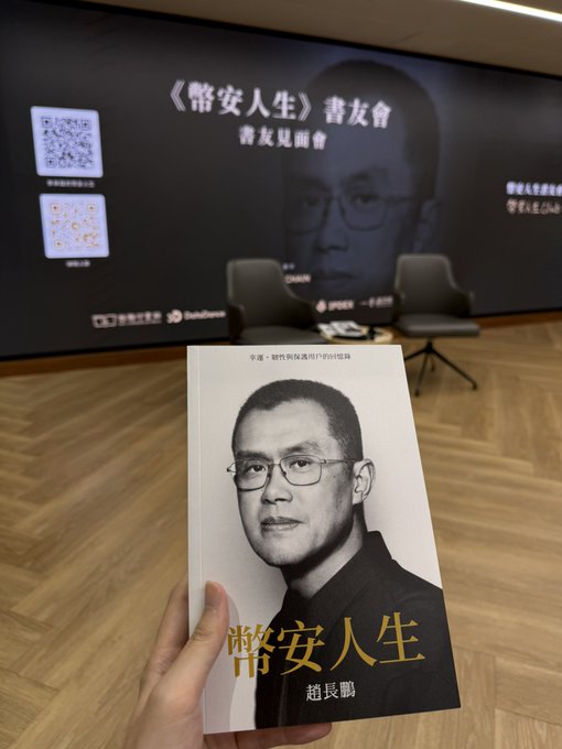 A person holds up a book titled "幣安人生" (Binance Life) by 趙長鵬 (CZ) at a book signing event, with a large screen in the background displaying the same title and "書友見面會" (Book Fan Meeting).
