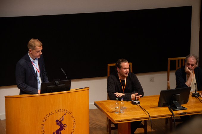 Three men participate in a panel discussion at a podium labeled 'Royal College of Surgeons of Edinburgh,' with one man speaking at the podium and two others seated at a table with laptops.