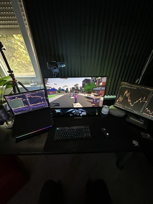 A multi-monitor trading setup on a black desk displays financial charts, a cryptocurrency price chart, and a video game window, complete with a keyboard, mouse, and microphone.