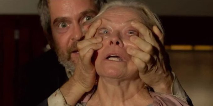 A horror movie still showing an older man with wide eyes grabbing the face of an older woman, who has a distressed expression.