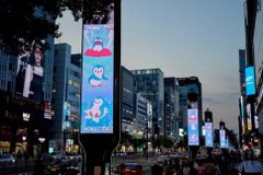 A city street at dusk featuring large illuminated billboards with cartoon characters, surrounded by tall buildings and cars on the road.