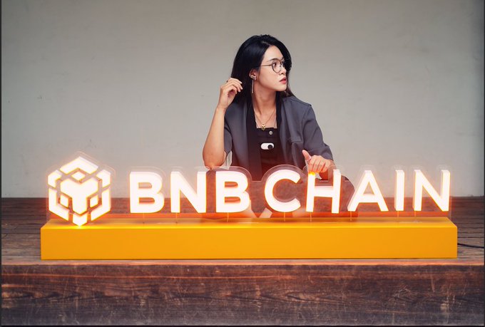 A woman with long dark hair and glasses sits behind a glowing 'BNB CHAIN' sign with an orange base and cube logo, wearing a dark blazer.