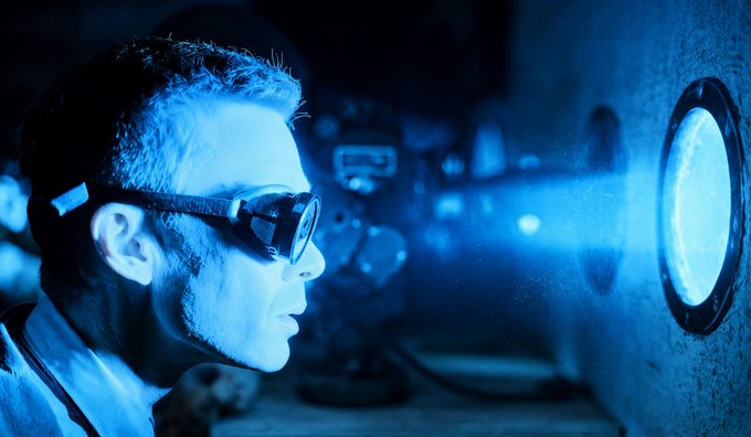 A cinematic, blue-tinted photograph showing a man wearing goggles in profile, staring intently at a glowing porthole or light source.