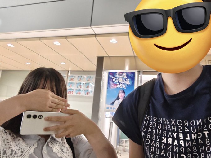 A person holds up a smartphone to take a selfie next to a friend whose face is covered by a sunglasses emoji, wearing a black t-shirt covered in mirrored letters that spell "CITY NIGHT SABBATH" and similar words. A poster in the background reads "第2弾 だいすきなあなたに" alongside a date of "5/21". The scene captures a candid, playful moment that matches the post's tone of lightly teasing a "ronin" friend's quirky fashion sense.