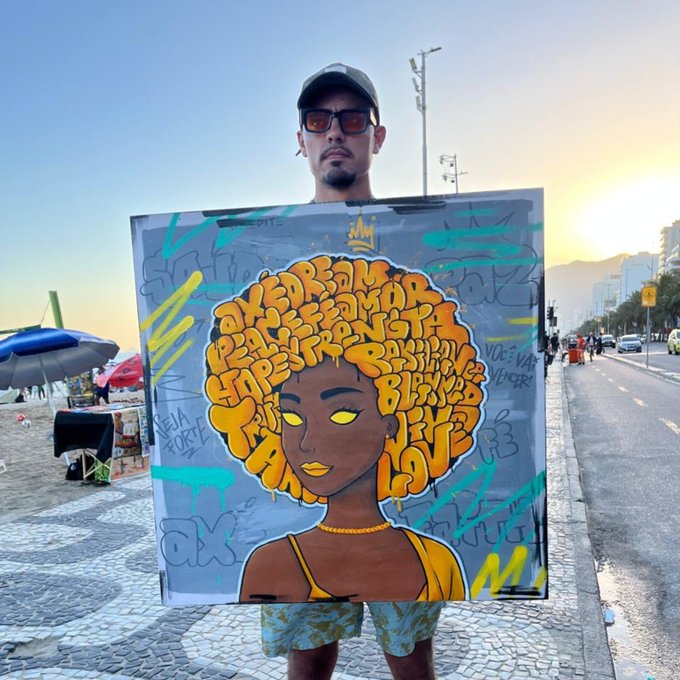 A man on a beach boardwalk at sunset holding up a graffiti-style painting of a woman with words in her hair.