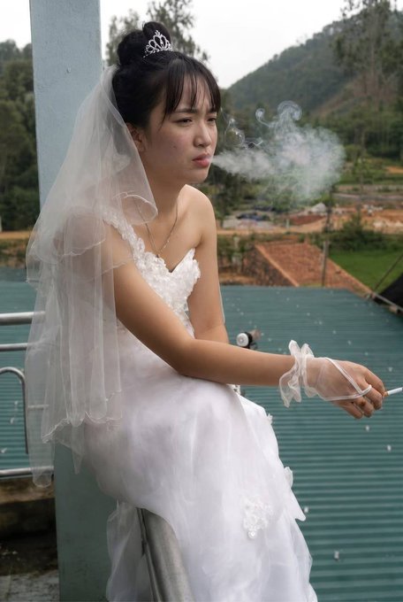 A young woman in a white wedding dress, veil, and tiara sitting on a rooftop while smoking a cigarette, with green mountains and a village in the background.
