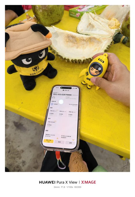 A photo taken with a 'HUAWEI Pura X View' shows a yellow table holding a BNB Chain plush toy, a smashed durian, and an Amoy figurine, with a smartphone displaying a BNB balance of '163.103.62879999'.