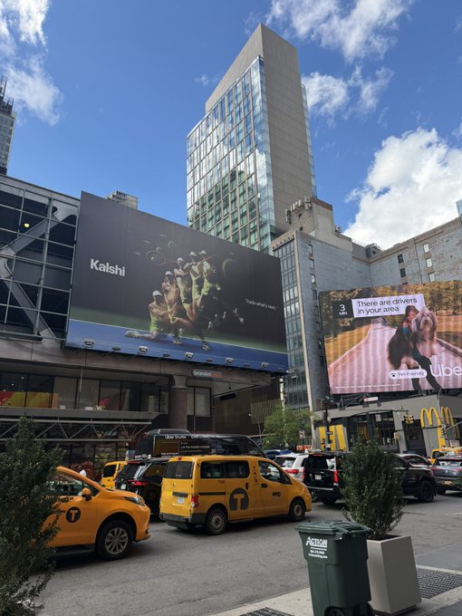 A New York City street scene featuring yellow taxis, a billboard advertising Kalshi with the text 'Trade what's next', and an Uber advertisement.