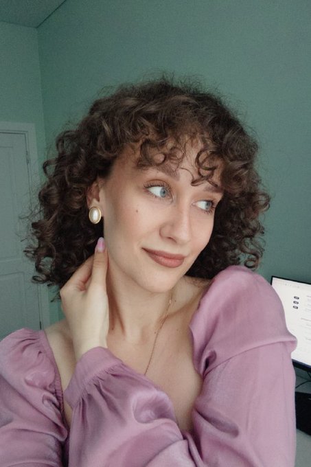 A young woman with curly brown hair and a mauve blouse poses thoughtfully against a teal wall, wearing pearl earrings and a delicate gold necklace, with sheet music visible in the background. Her knowing, sidelong glance and subtle smile cleverly capture the caption's sarcastic tone about feigning concern over the Clarity Act failing to pass.