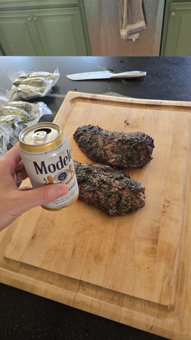 A hand holds a can of Modelo beer next to two cooked pieces of flank steak resting on a wooden cutting board.