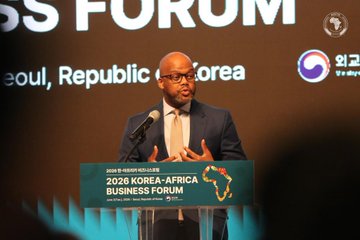 In the image, there is a man standing behind a podium giving a speech at an event with a screen behind him displaying text related to the "The AfCFTA market (1.4B people) is transforming into a strategic partnership with Korea! Protocols for digital trade & advanced tech like IOTA create secure supply chains & new opportunities for e-commerce, automotive & pharma." The man appears confident and knowledgeable as he speaks.