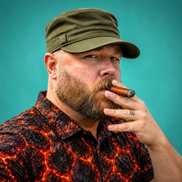 A bearded man wearing an olive-green military-style cap and a dark red shirt with a lava-like crack pattern smokes a thick cigar against a teal background.