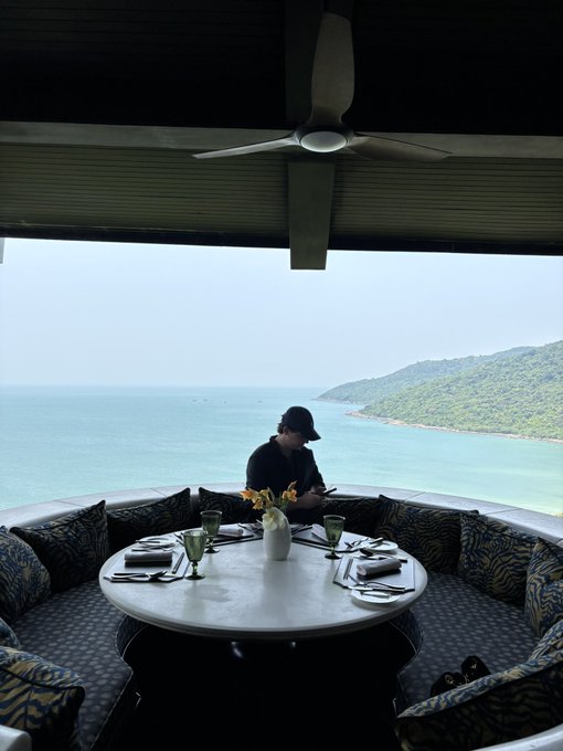 A person wearing a black cap sits at a circular dining table set for a meal, framed by a large window offering a scenic view of a turquoise sea and green hills.