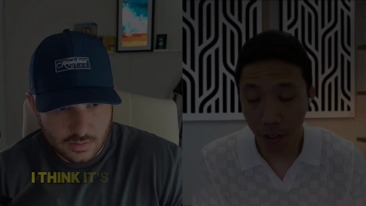 The image shows two men wearing baseball caps sitting next to each other, with one man looking at a computer screen while the other looks away from the camera. The background features a white wall adorned with black and white geometric patterns.