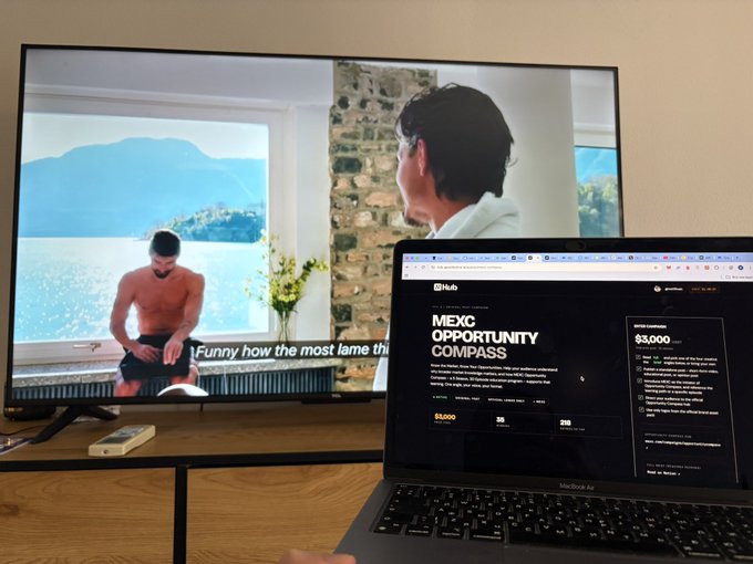 A laptop displaying a 'MEXC OPPORTUNITY COMPASS' webpage next to a TV screen showing a video of a shirtless man.