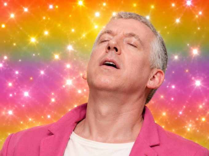 A man with gray hair wearing a bright pink blazer and white t-shirt poses with his head tilted back, mouth open, and eyes closed against a sparkling rainbow-colored background.