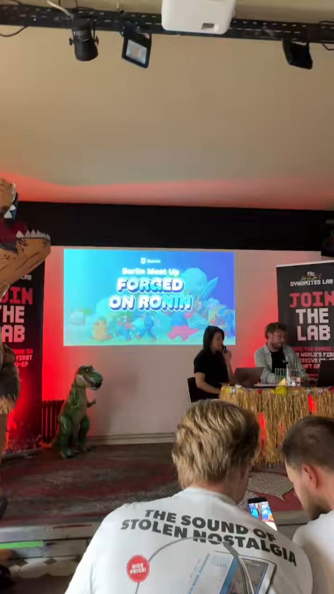 The image shows a group of people gathered around a stage, with one man sitting in front of a large screen displaying "Forced On Rounded" from the movie Toy Story. The audience appears engaged as they watch the presentation or event taking place on the stage.