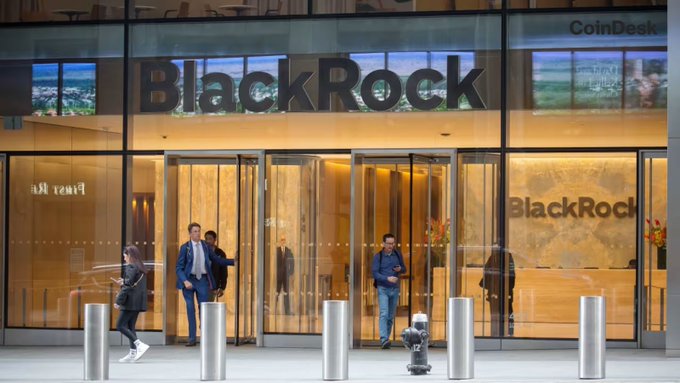 The entrance to a BlackRock building with people entering and exiting through the glass doors, with the company's logo displayed above and a CoinDesk watermark in the corner.
