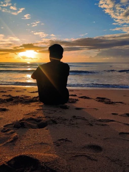 A silhouette of a person sitting on a sandy beach at sunset, with footprints in the sand and waves rolling onto the shore under a partly cloudy sky.