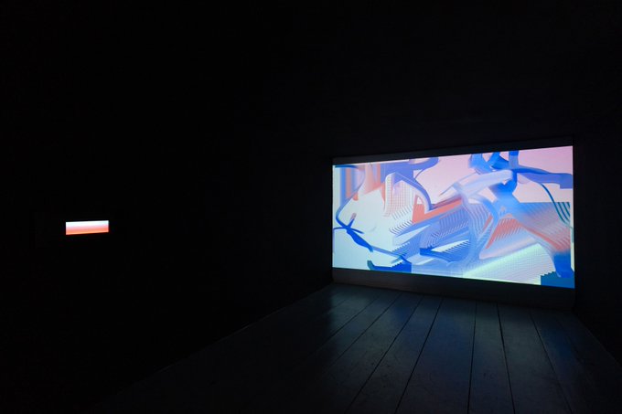 The image shows a dark room with a large screen displaying a colorful abstract painting, possibly a digital art piece or a projection of an artwork. The room appears to be a gallery space where artists showcase their work. The painting's vibrant colors contrast against the black background, making it stand out prominently in the scene.
