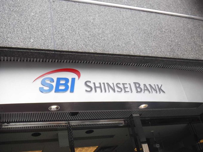 The image shows an SBI Shinsei Bank sign outside a building. The sign displays the bank's logo and name along with information about their pilot project for exchanging digital currency, such as Bitcoin or Ethereum. This pilot project aims to provide customers with the option to convert their deposit interest into digital currency, which can be used in various online platforms.