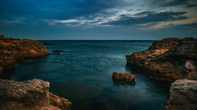 A moody, twilight photograph of a rocky coastline with calm, deep blue water framed by dark stone cliffs under a cloudy sky.