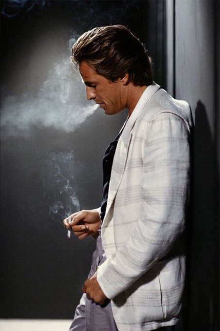 A man in a light-colored plaid blazer stands in profile, smoking a cigarette with smoke curling upward against a dark background.