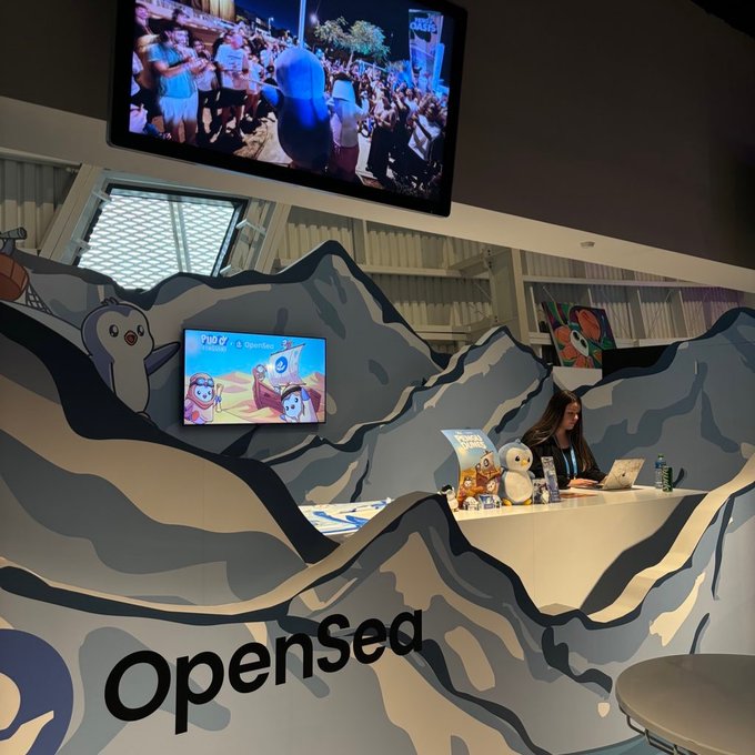 A woman works at an OpenSea booth styled with icy mountain displays, Pudgy Penguins imagery, and a "Play" screen featuring NFT collections, alongside a TV showing a crowded event—reflecting her role in community events and brand growth at Pudgy Penguins.