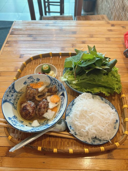 A meal served on a woven bamboo tray includes a bowl of broth with meatballs and vegetables, a plate of white rice noodles, and fresh green herbs.