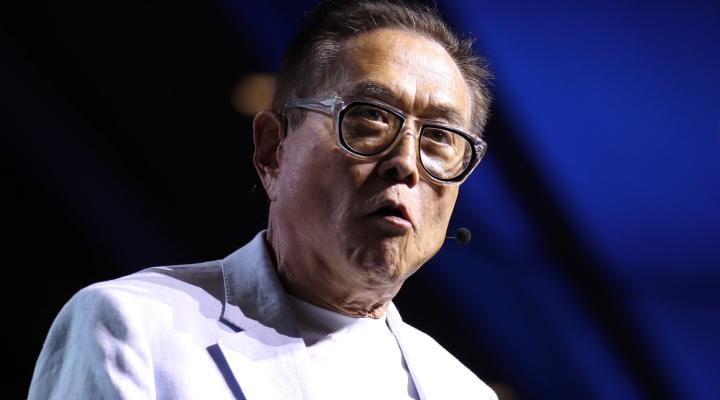 The image shows Robert Kiyosaki wearing glasses and a headset microphone, speaking at what appears to be a stage event with a blue-lit background. The caption relates to the post about his rumored statement regarding XRP potentially making investors wealthy, though his expression appears serious as he addresses an audience.