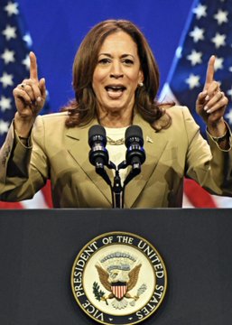 Kamala Harris stands at a podium bearing the Vice President of the United States seal, speaking with both index fingers pointed upward against a backdrop of American flags. The image accompanies news of her upcoming campaign appearance alongside Michigan Senate nominee Abdul El-Sayed.
