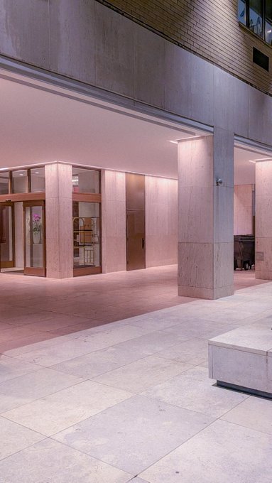 An empty outdoor plaza features light-colored stone flooring and pinkish stone columns and walls under warm lighting at dusk or dawn.