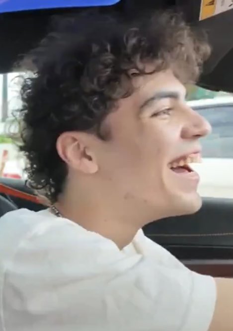 Close-up of a young man with curly hair laughing in profile while sitting in a vehicle.