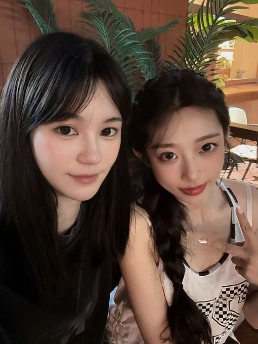 A close-up selfie of two young women posing together indoors, one with long dark hair and the other with a braid making a peace sign.