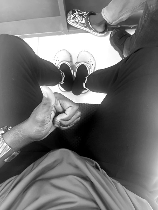 A black-and-white photo taken from above shows a person sitting with clenched fists on their lap, wearing a watch, with another person's feet in white Crocs visible nearby.
