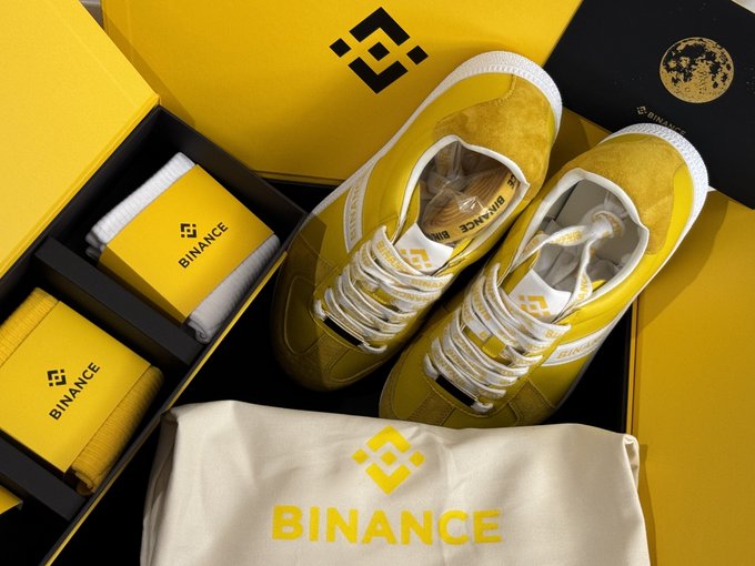 A pair of yellow suede sneakers with white laces and soles sits alongside Binance-branded yellow packaging boxes and a cream drawstring bag with the Binance logo.