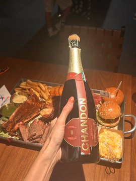 A hand holds up a bottle of Taittinger "Prestige Rosé" Brut Champagne in front of a large tray loaded with fries, sliders, and assorted meats, celebrating a small $BNB crypto profit. The casual feast captures the relaxed weekend vibe of toasting a modest trading win.