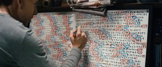 Man writing or marking numbers on a large sheet of paper covered with handwritten digits in red and blue, likely lottery or statistics tracking.