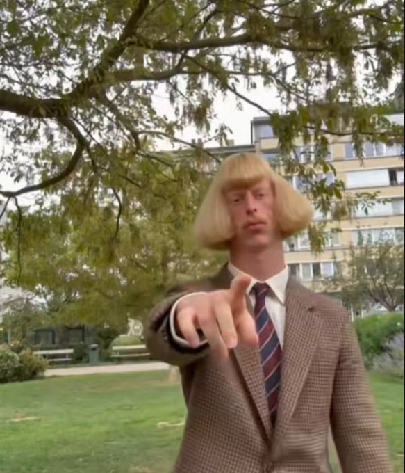 A man with a distinctive blonde bowl haircut, wearing a brown tweed jacket and striped tie, points directly at the camera in a park setting.