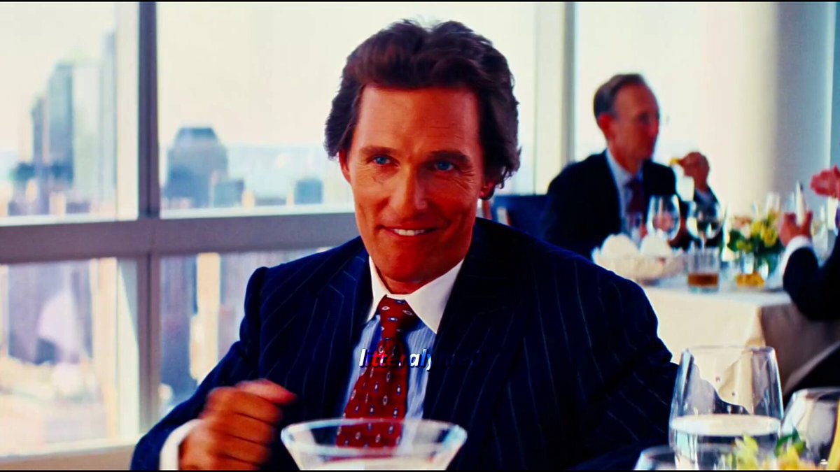 The image shows a man sitting at a table with wine glasses and plates of food. He is wearing a suit and tie, giving off an air of sophistication or formality. The background reveals that he is dining alone, possibly in a restaurant setting. There are also other people present in the scene but they are not the main focus of the image.