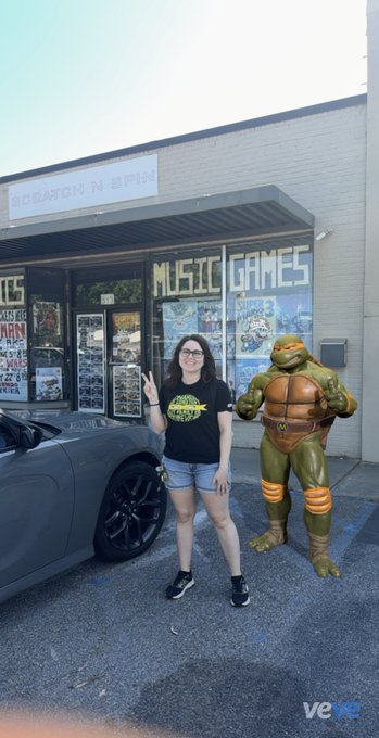 A photo of a woman in a Teenage Mutant Ninja Turtles t-shirt making a peace sign next to a person in a Michelangelo costume outside a 'MUSIC GAMES' store, with a gray car parked nearby.