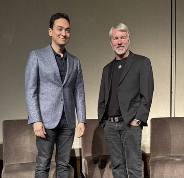 Two men pose together indoors, one in a blue blazer and the other in a black suit jacket with a beard.